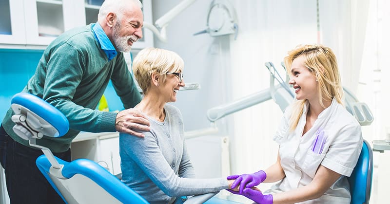 An older couple in a dental room speak to a nurse with purple gloves