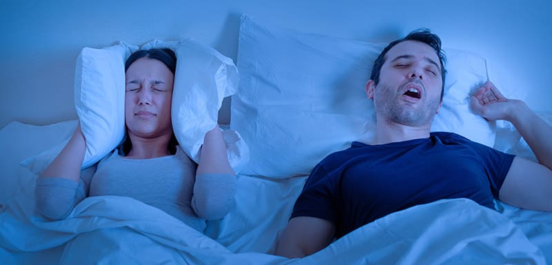 A woman holding a pillow to her ears as her husband sleeps on a pillow with an open mouth