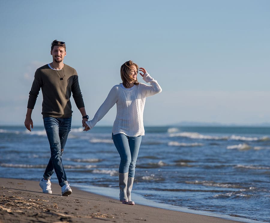 A couple in jeans and long sleeve shirts walk along a beach under the sunny sku