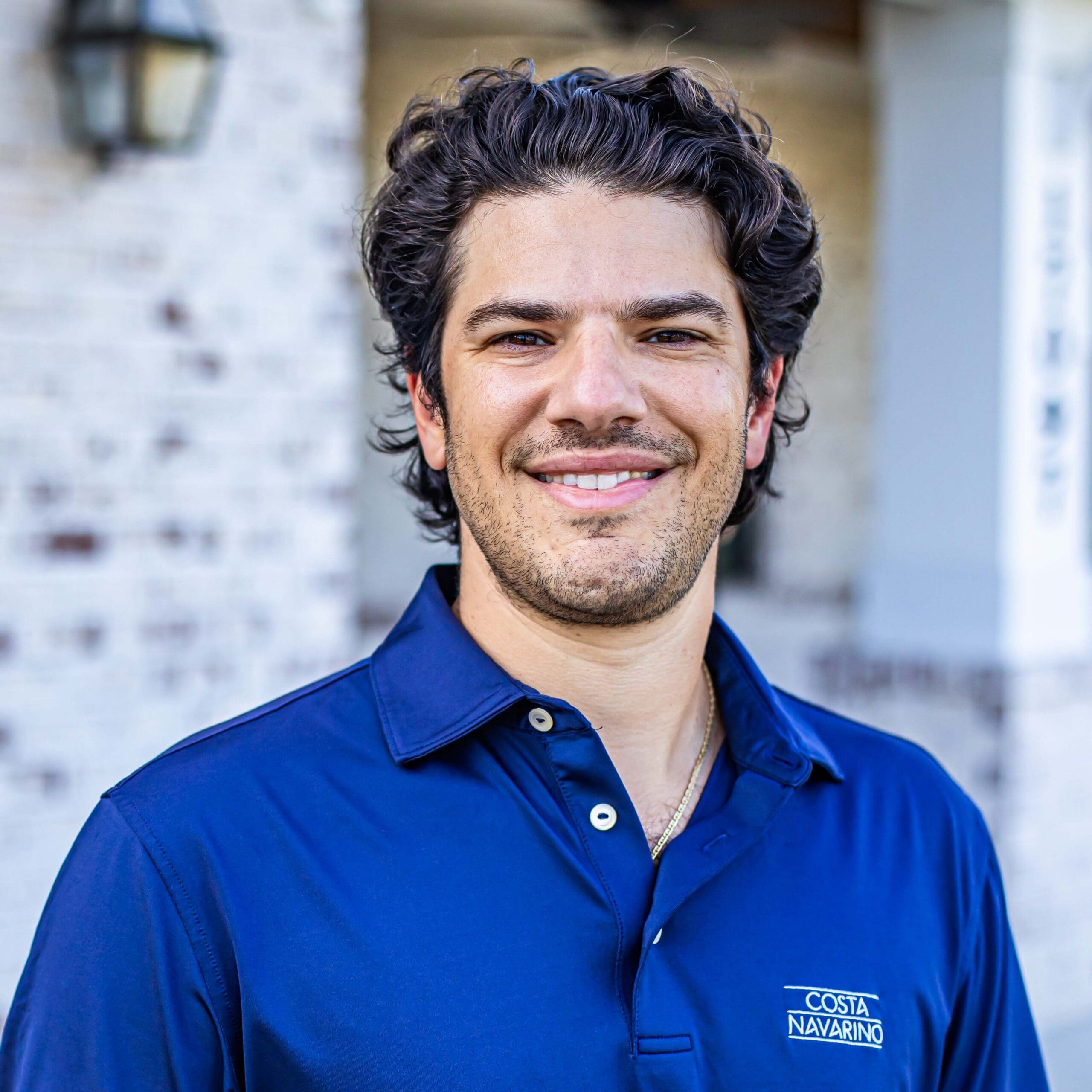 Dr. Andrew Troupes smiling in front of a white brick wall and wearing a gray polo