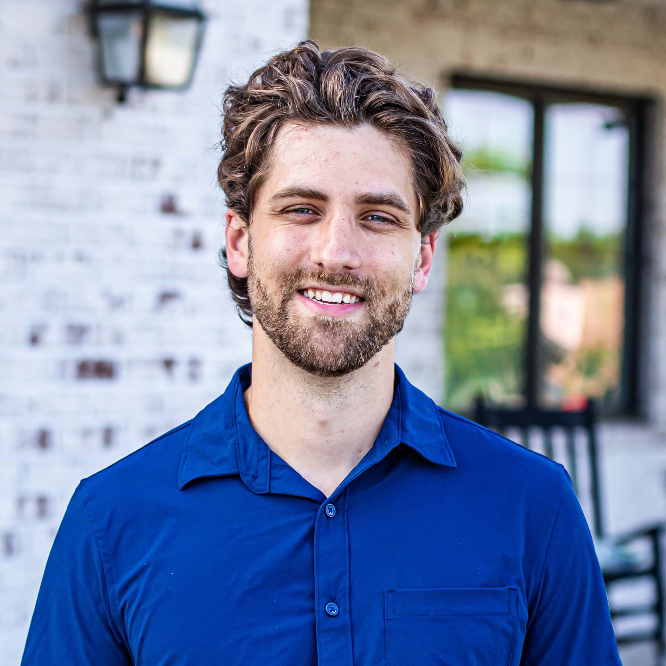 Dr. Mark Castor smiling in a blue dress shirt standing in front of a palm tree